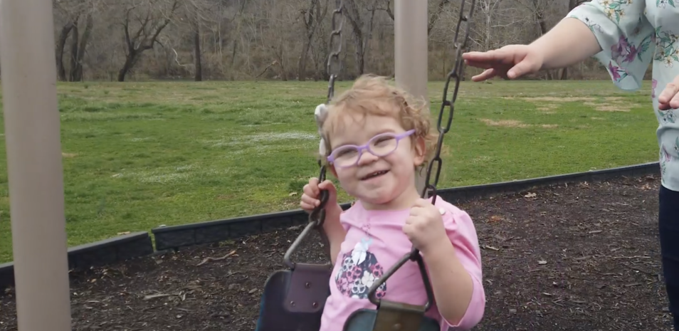 A toddler wearing glasses and a pink shirt being pushed by her mother on a swing set.