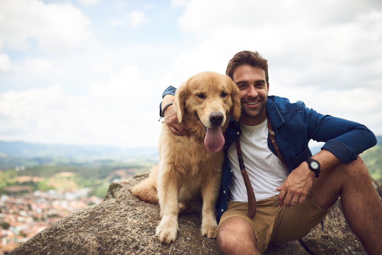 Man n blue jacket, white shirt, and tan shorts hugs a panting golden retriever while sitting on a rocky ledge overlooking a town