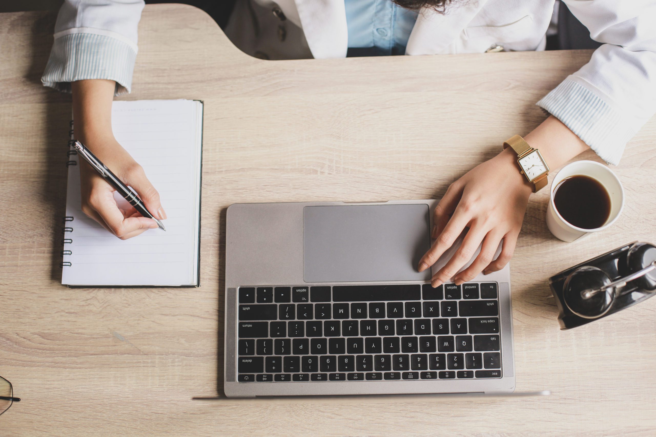 Aerial shot of woman working at a desk. One hand is writing on a notepad, the other is resting on a laptop beside an alarm clock and a cup of coffee.