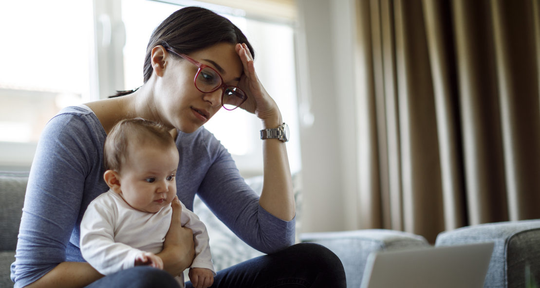 Brunette woman in red glasses holds a baby in her lap and looks tiredly at a laptop.