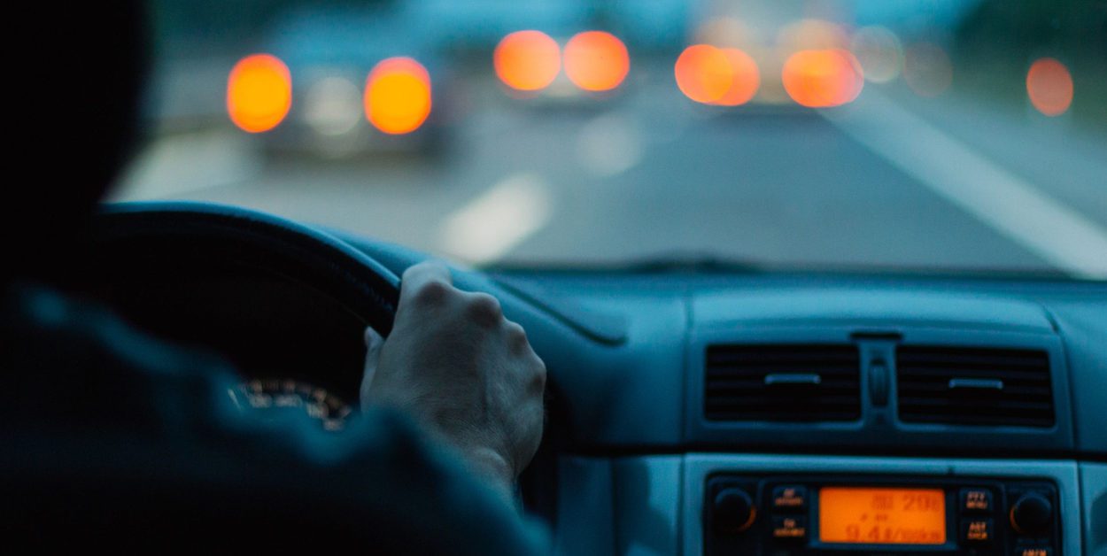 Close up of a driver's right hand on a steering wheel and the car's radio and the headlights ahead glowing orange