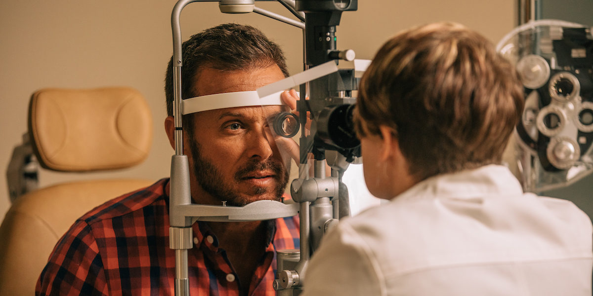 Dark-haired male patient in a red and blue flannel undergoes an eye exam with a short-haired doctor who is facing away from the camera