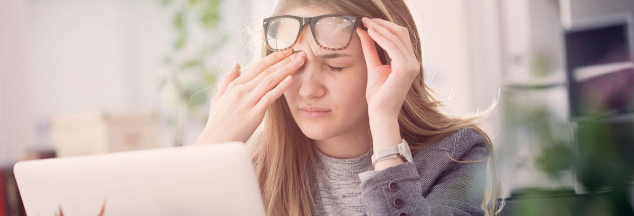 Young woman sitting at her laptop lifts her glasses up to her forehead and rubs her eyes in a tired way