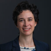 Headshot of woman with short dark hair in a navy blazer in front of a dark background.